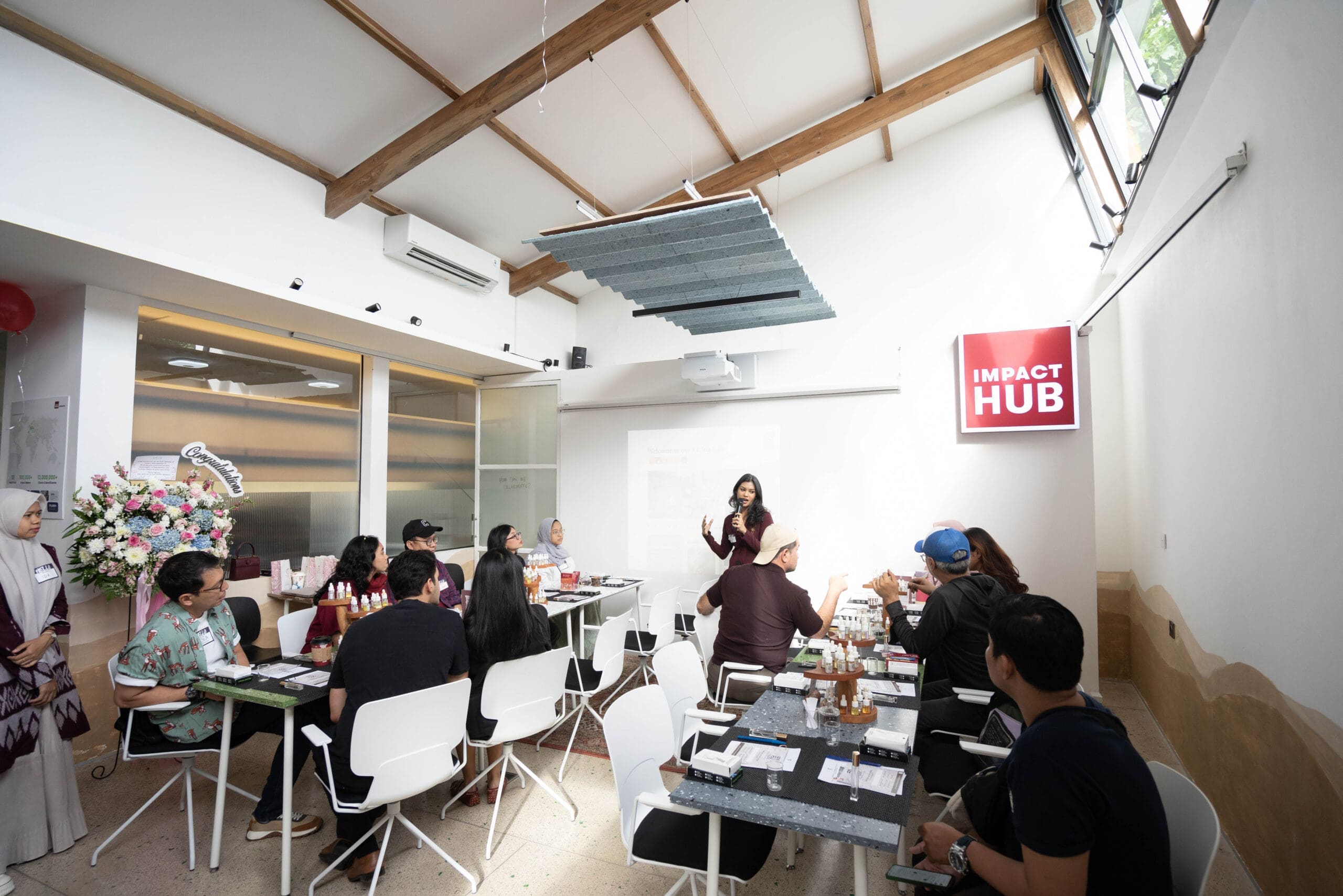 A speaker presenting to an audience seated at tables inside Impact Hub Jakarta, with the Impact Hub logo on the wall and people listening during a program session at the grand opening event.