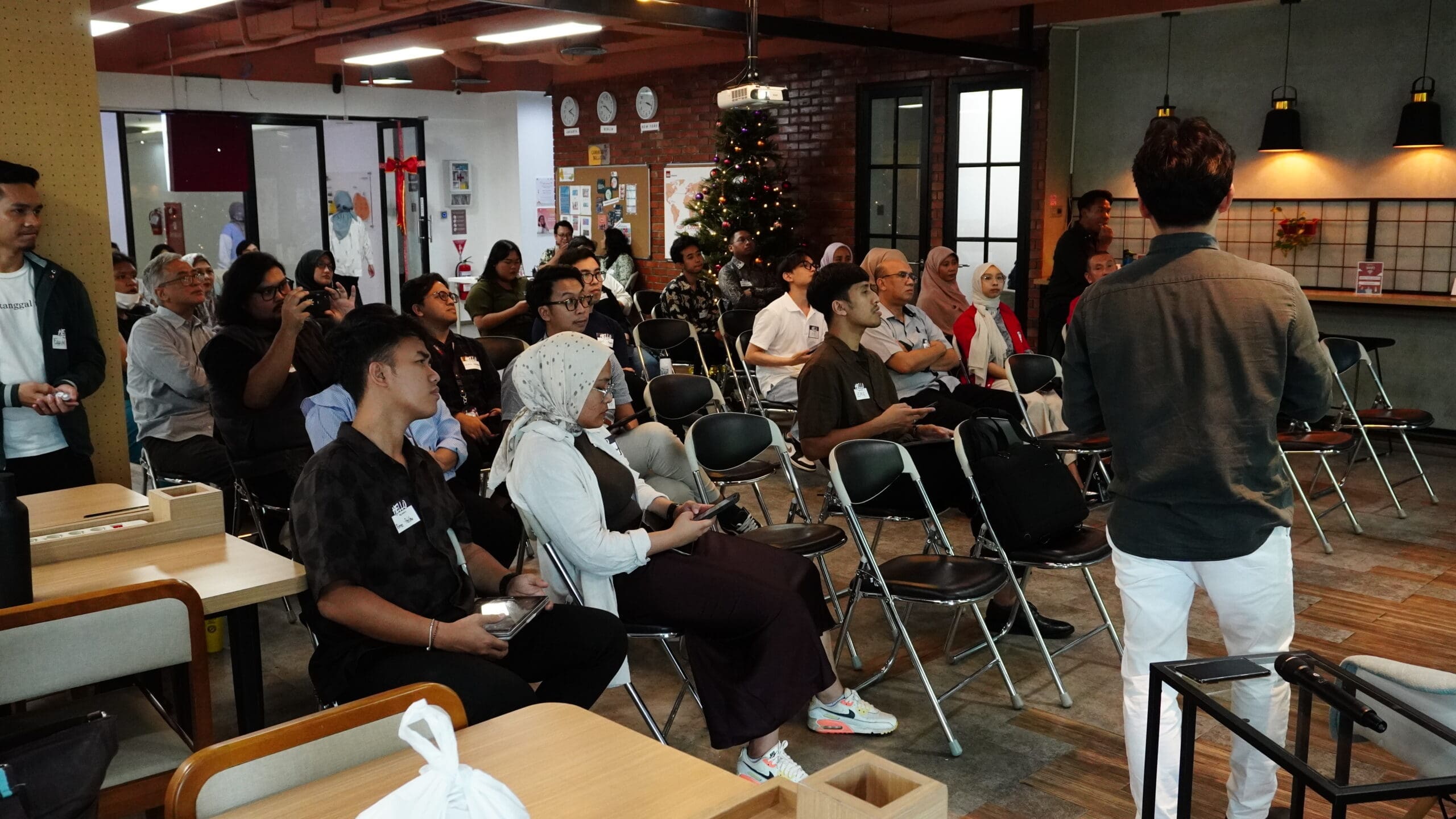 Participants seated in rows at the Embedding Circularity Toolkit launch event, listening attentively to a speaker presenting at the front of the room. The audience is diverse and engaged, some taking notes on their phones, inside a warm, modern community space with brick walls, folding chairs, and a decorated Christmas tree in the background, capturing a focused and collaborative learning atmosphere.