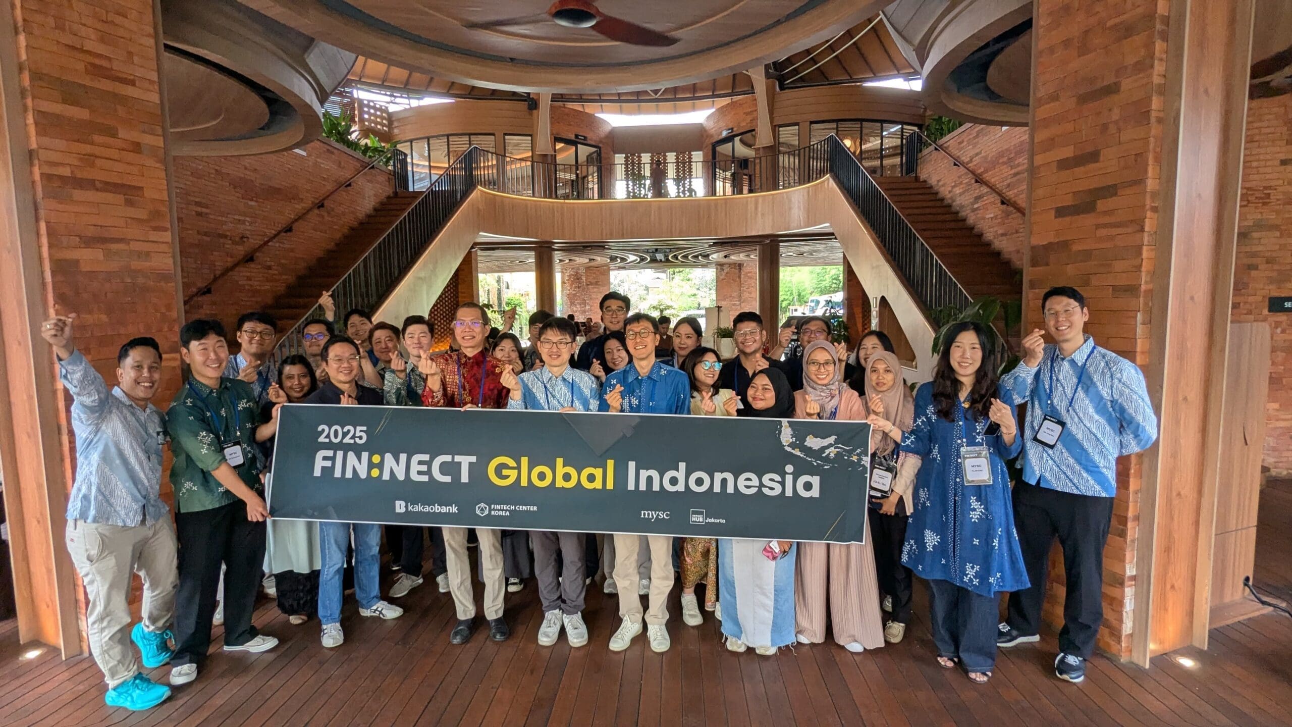 A large group of participants posing together indoors at the 2025 FIN:NECT Global Indonesia event, holding a banner that reads “FIN:NECT Global Indonesia.” The group smiles and makes celebratory hand gestures, wearing batik and business-casual outfits. They stand in a spacious venue with warm wooden floors, brick walls, and a grand staircase in the background, capturing a moment of cross-border collaboration between MYSC and Impact Hub Jakarta.
